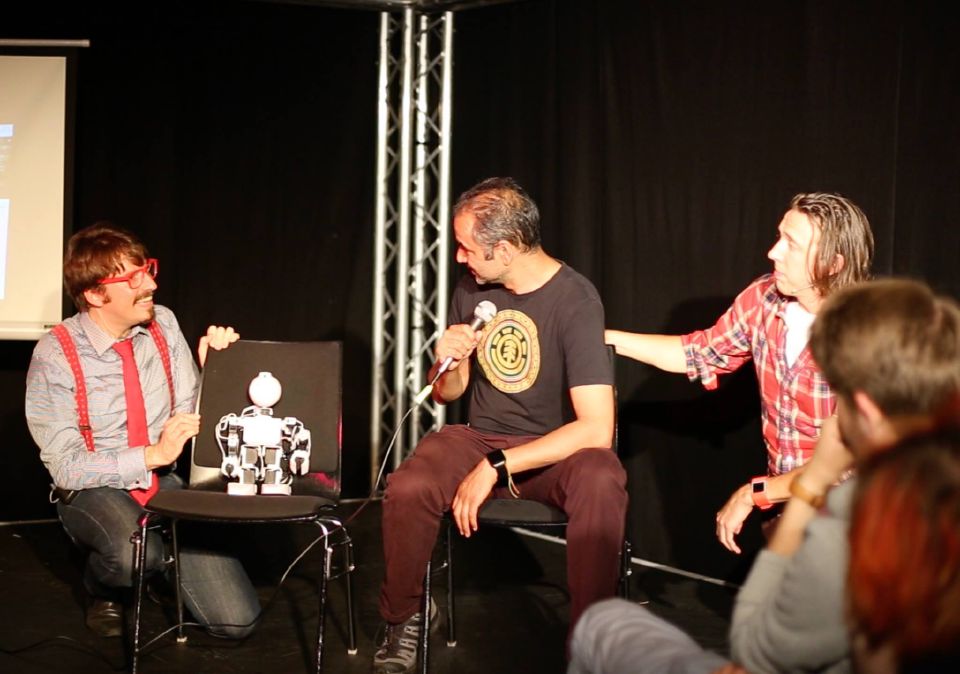 Two improvisers on stage with a small humanoid robot on a chair, beside an audience volunteer holding a microphone.
